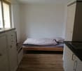 Single bedroom with bed, wooden floor, white walls, two windows, built-in closet, and open kitchen.