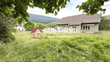 House with a brown roof, two windows, and a white wall, situated in a grassy area. A watermark from 'SAINT-IMMO' is displayed in the center of the image, with another house in the background and a mountain in the distance. The sky is clear and blue.