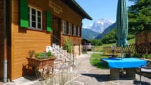Wooden chalet-style building with green shutters, surrounded by mountains and greenery. Outdoor seating area with tables and chairs, and a covered patio. Parking space visible in the foreground.