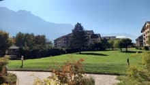 A wide grassy area with well-maintained trees and bushes, leading to a building complex with apartments in the background. Mountains can be seen in the distance.