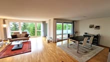 Spacious open-plan living and dining area with large windows overlooking a balcony or terrace. The room features hardwood floors, a brown leather sofa, a coffee table, and a dining table with chairs. There is also a media console and shelving unit against
