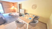 The image shows a combined living room and dining area. The living room has a wooden accent wall, a round decorative wall hanging, and a coffee table. The dining area has a wooden table with four gray upholstered chairs, and a sideboard unit. The room has