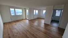 Spacious open-plan living area with large windows, hardwood flooring, and a view of the city outside. The room appears to be a living room or combined living/dining area.