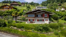 Detached house, wooden exterior, nestled in a hilly area, surrounded by greenery, multiple windows, driveway leading to the entrance.