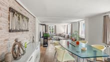 A spacious living room with brick wall, white painted walls, a sofa, coffee table, glass table, and chairs. It has a fireplace and white furniture. There is also a brick chimney, a white cabinet, and a glass top table.