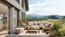 Spacious outdoor patio with wooden deck, comfortable seating area with sofa and armchairs, coffee table, decorative plants and flowers, and scenic mountain view in the background