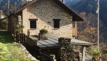 a stone house with a wooden front porch, elevated ground floor, chimney, windows, flag, and lush greenery, nestled against a mountainous backdrop