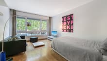 A room with a bed, a couch, a TV, a lamp, a table, and a window with a view of the outside.
