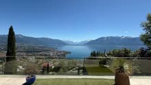 Outdoor terrace with a view of the lake and mountains, glass railing, potted plants, green grass