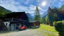 2 story wooden house, gravel driveway, steps leading to porch, red car, small wooden cabin, sunny weather, mountains in the background
