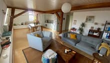 Open living room with wooden floor, wooden beams, white walls, two sofas, wooden coffee table, two armchairs, carpet, dining table, chairs, kitchen, microwave, refrigerator, potted plants, shelves, decorative items