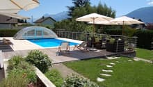 Outdoor swimming pool surrounded by patio furniture and umbrellas, with a grassy lawn and landscaping visible.