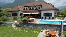 Large two-story house with a tiled roof, surrounded by mountains. The house has a patio with a swimming pool, wooden furniture, and a stone wall. There are also parking spaces on the property.