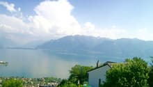 The image shows a scenic view of a lake surrounded by mountains and lush greenery. The lake appears to be calm and reflective, with buildings and houses visible along the shoreline. The sky is clear with some fluffy white clouds, creating a picturesque an