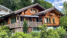 Two-story wooden chalet-style house with a balcony, surrounded by lush greenery and trees