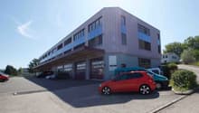 3-story commercial building with a modern, angular design featuring purple/gray siding and large windows. There is a parking lot in front of the building with several cars parked.