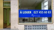 Exterior of a commercial building, white and gold concrete walls, glass entrance with brass frame, blue sign indicating 'A LOUER' with contact number.