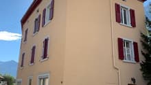 A beige building with 7 windows, red shutters, white accents and a red roof