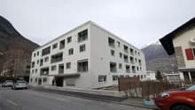 A multi-story apartment building with a modern, minimalist design. The building has several balconies and large windows. The exterior is painted in a light gray color. There are cars parked in front of the building, and the surrounding area appears to be