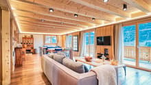 Open-plan living area with wooden beams, large sofa, center table, wooden floors, balcony, large windows