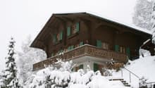 2-story wooden chalet-style house with a balcony, covered in snow in a winter landscape