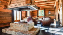 Rustic log cabin interior with wooden walls and ceiling, stone fireplace, leather armchairs, and a coffee table on a tiled floor