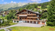 Large wooden chalet, multiple balconies, garage, set in a mountainous area with green slopes and pine trees.