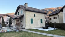 Detached house, two stories, stone facade, green windows, driveway, surrounded by grass and bushes, mountain view