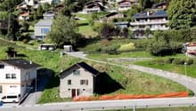 Aerial view of a hillside with several residential properties. Two-story houses with multiple windows. A grey detached house with balconies is under construction. Parking spaces are located near some of the houses. A pathway and a road with a guardrail ar
