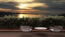 The image shows an outdoor patio or terrace overlooking a scenic lake or body of water. The patio has a round table and two chairs, providing a comfortable seating area to enjoy the beautiful sunset view over the water. The landscape is filled with lush g