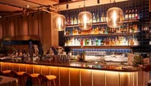 Well-stocked bar with a variety of liquor bottles, glassware, and large pendant lights hanging above the bar counter. The bar has a modern, cozy atmosphere with wooden accents and tiled walls.