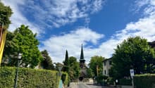The image shows a picturesque street in a small town or village. The scene features a church steeple with a green roof, surrounded by lush green trees and hedges. The street is lined with buildings and has a parking area visible. The sky is blue with fluf