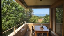 Covered balcony with wooden floor, glass railing, and a dining table with chairs overlooking a scenic view of trees and distant buildings