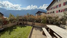 Garden: wooden table, benches, gravel path, green lawn, bushes, fireplace, wooden deck, mountains in the background.