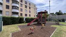 Playground with slide, climbing structure, and horsey ride toys, bordered by bushes, adjacent to a building with balconies.