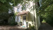 A two-story house with a yellow exterior and a brown roof. The ground floor is raised with concrete steps leading to the entrance. The windows are adorned with white shutters. The house is surrounded by a yard with trees and shrubs. There is a pathway lea