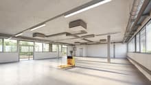 Large open commercial space with concrete floors, exposed ceiling, and large windows. The space is equipped with overhead lighting fixtures and a yellow pallet jack in the center of the room.