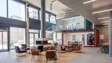 Large open-concept office space with concrete walls and ceiling, large windows, leather armchairs and sofas, and a spiral staircase leading to a mezzanine level.