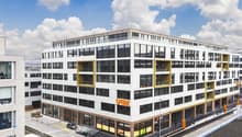 A modern, multi-story commercial building with a large glass facade, yellow accents, and a parking area in front.