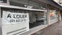 Shopfront with glass walls and doors, blue awning, white exterior, parked cars outside, signboard with a phone number and 'A LOUER' written in black