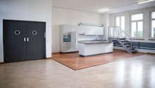 Spacious open-plan kitchen with white cabinets, built-in appliances, and hardwood flooring. Large windows provide natural light. Metal staircase visible in the background.