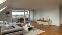 A modern and open living room with large windows and sliding glass doors leading to a balcony with an outdoor dining area and stunning views of the forest. The room features wooden floors, a comfortable grey sofa, a white coffee table, and decorative item