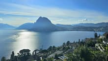 Lake Lugano with mountains in the background, a clear blue sky, town in the distance, greenery