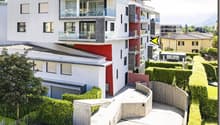 Modern apartment building with multiple floors, white and red facade, glass balconies, fenced entrance, hedges, and a small garden
