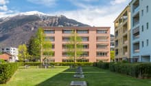 Modern apartment building with multiple floors, surrounded by greenery and a playground, with a view of the mountains