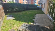 The image shows a residential apartment complex with several multi-story buildings. The buildings have a mix of orange and gray colors. There is a small grassy area with a palm tree in the foreground, and a paved walkway leading to the buildings. The comp