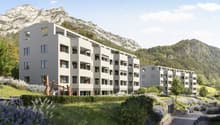 Modern apartment complex on a slope with large windows and balconies. Surrounded by greenery and a car park.