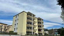 Multi-story apartment building with balconies, beige exterior, and surrounded by trees and greenery.