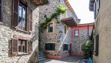 This image shows a traditional stone building with wooden shutters and balconies. The building has a rustic, Mediterranean-style architecture with a cobblestone pathway leading to the entrance. There are potted plants and a small bench visible, adding to