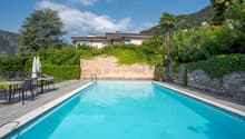 Two-story house with a swimming pool, surrounded by lush greenery and palm trees. The house has a balcony or terrace overlooking the pool area.